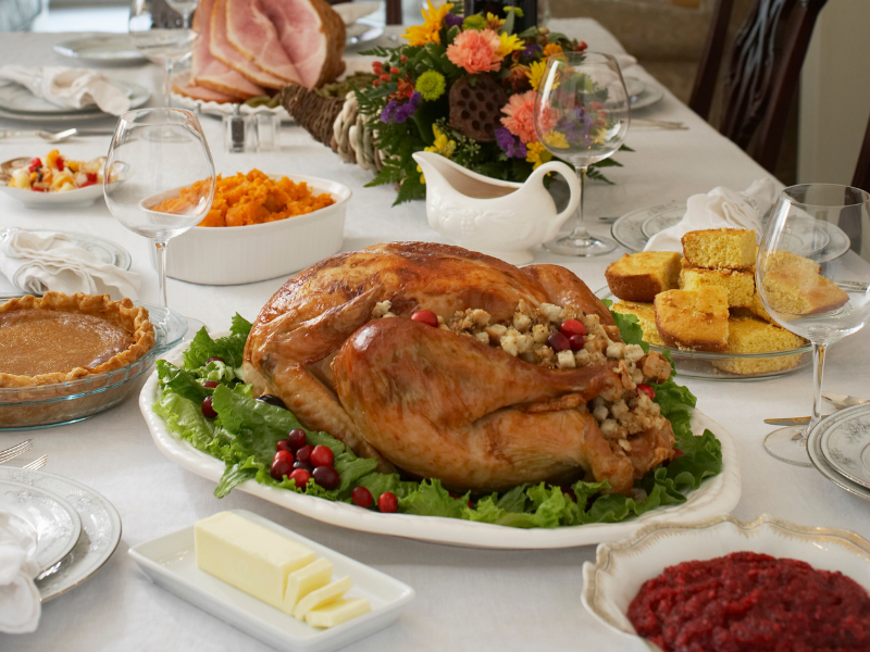 picture of a table with a white tablecloth with a cooked tukey, cornbread, pumlkin piem carnberry sauce and ofhter thanksgiving sides,. There is also a bouquet on the table and empty wine glasses. 
